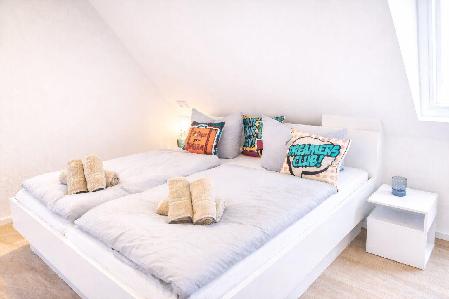 Schlafzimmer mit Doppelbett, Kissen und Handtüchern. Fenster und Nachttisch sind sichtbar.