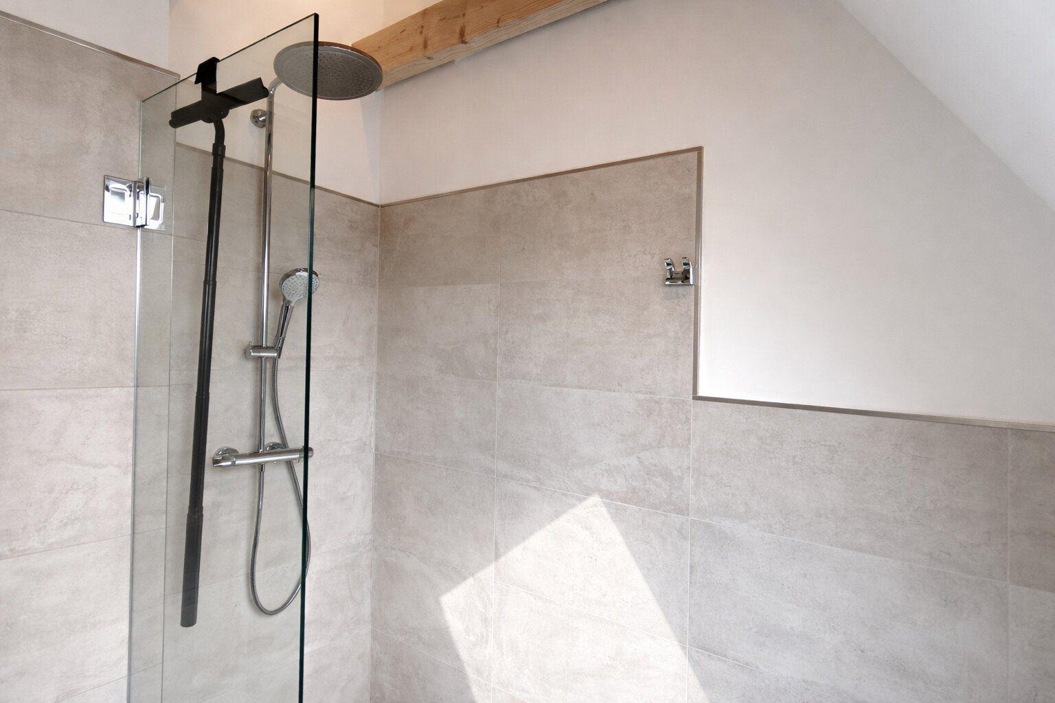 Dusche mit Glaswand, Duschbrause und Handbrause. Wandfliesen und Holzbalken sichtbar.