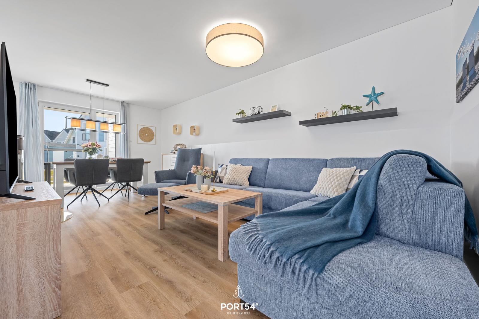 Wohnzimmer mit blauem Sofa, Holztisch und Fernseher. Hintergrund zeigt Essbereich mit Tisch und Stühlen.