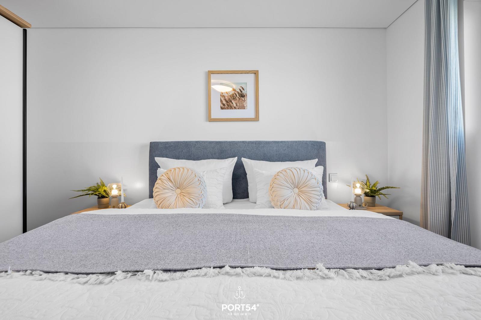 Schlafzimmer mit Bett, Kopfkissen und Nachttischen. Wandbild und Vorhänge sind sichtbar.