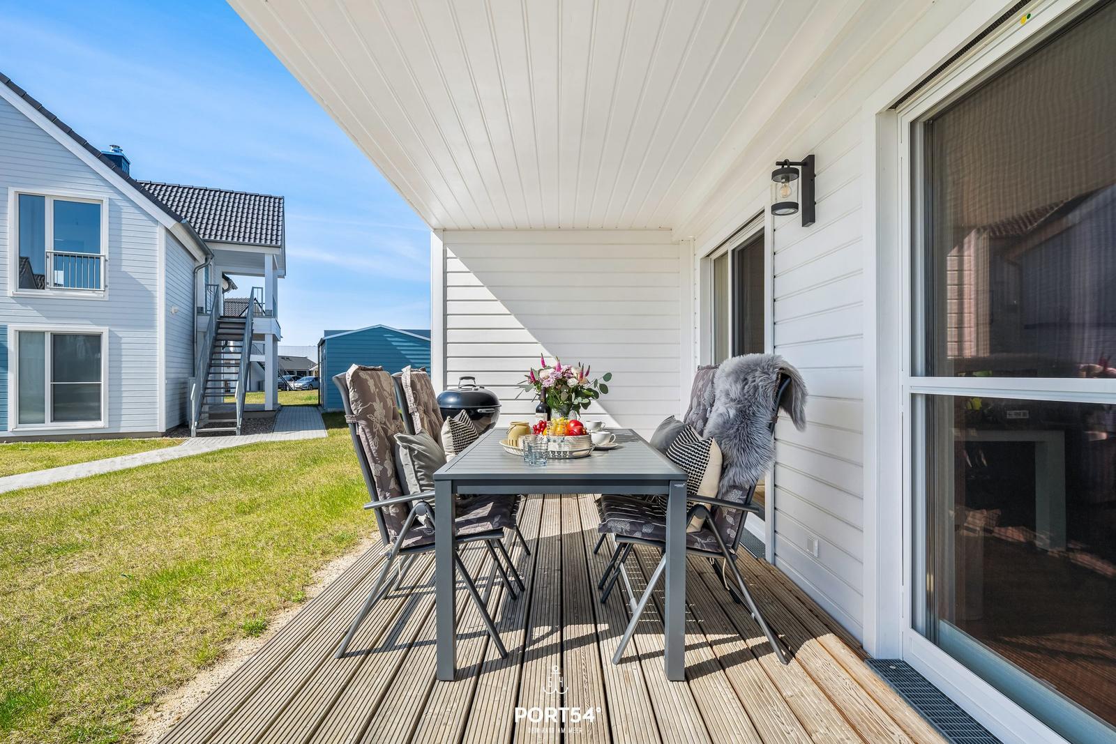 Terrasse mit Tisch, Stühlen und Grill. Hintergrund: Haus und Grasfläche.