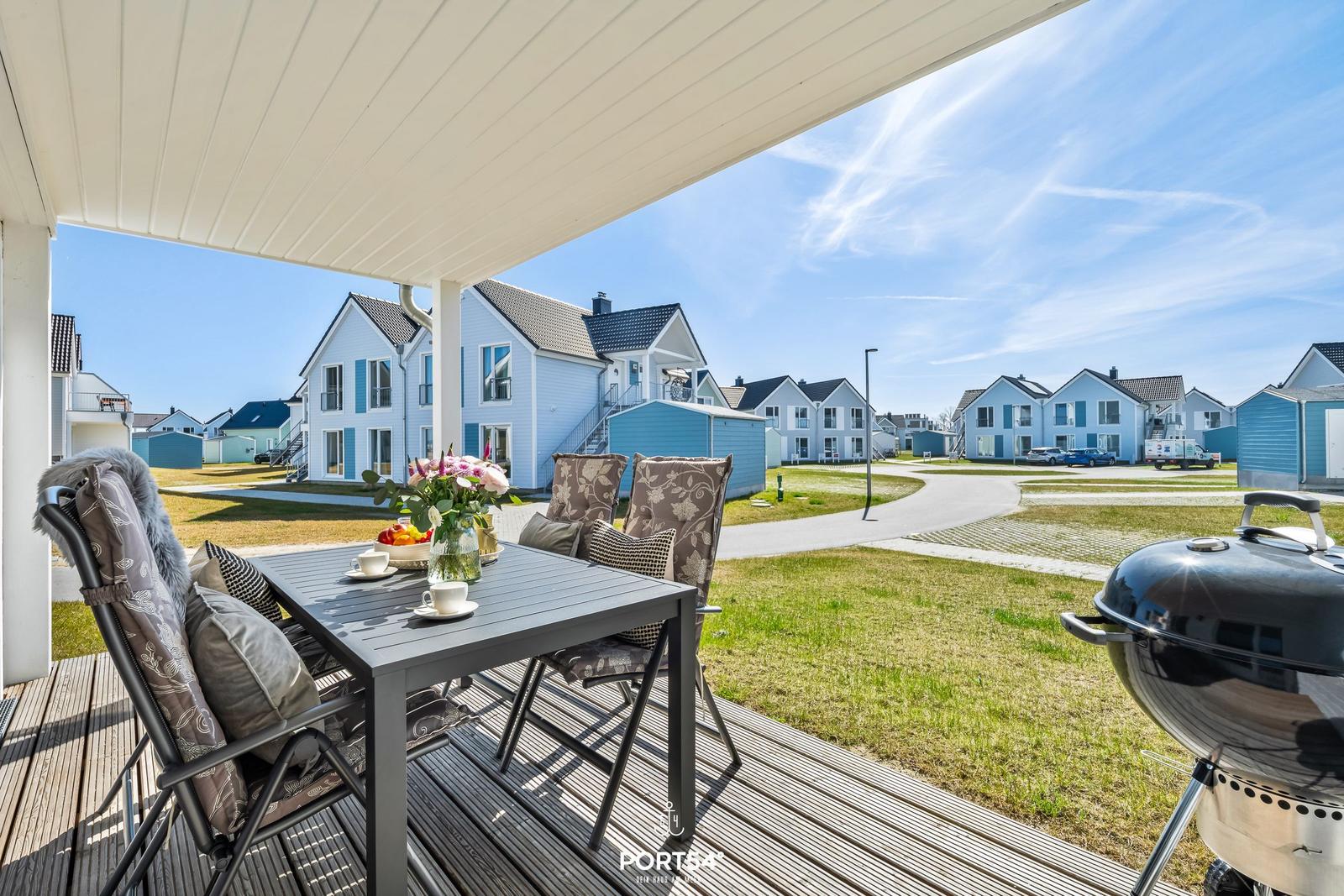 Terrasse mit Tisch, Stühlen und Grill. Blick auf Wohnhäuser und Grünflächen.