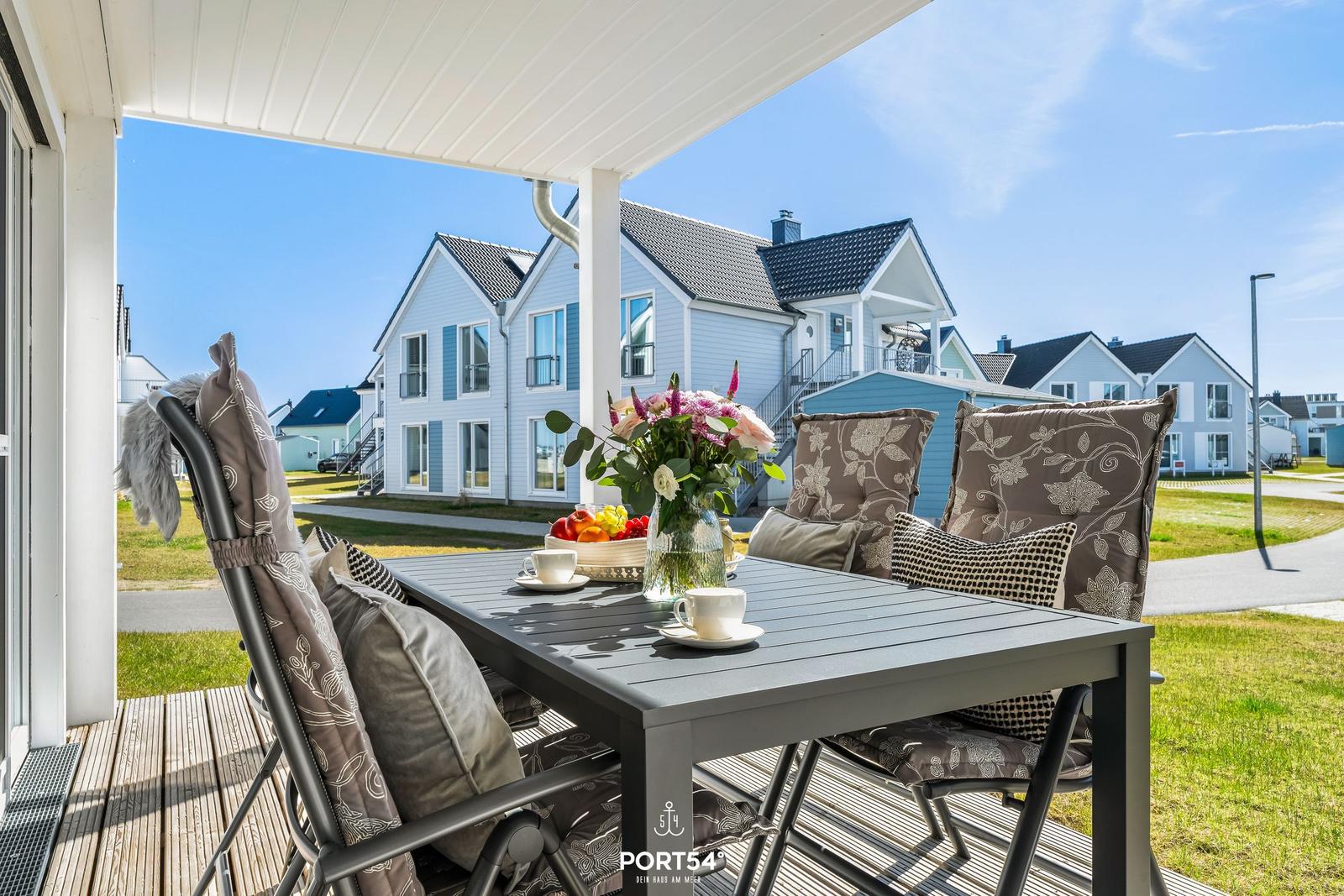 Terrasse mit Tisch, Stühlen und Blumen. Im Hintergrund sind Häuser zu sehen.