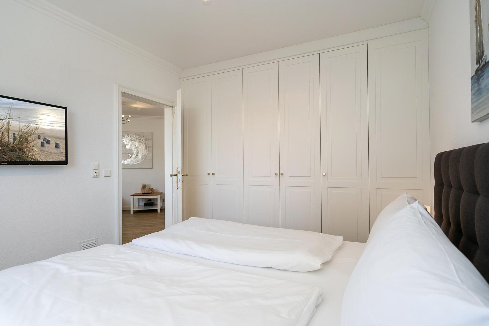 Schlafzimmer mit weißem Bett, Kleiderschrank und Fernseher an der Wand.