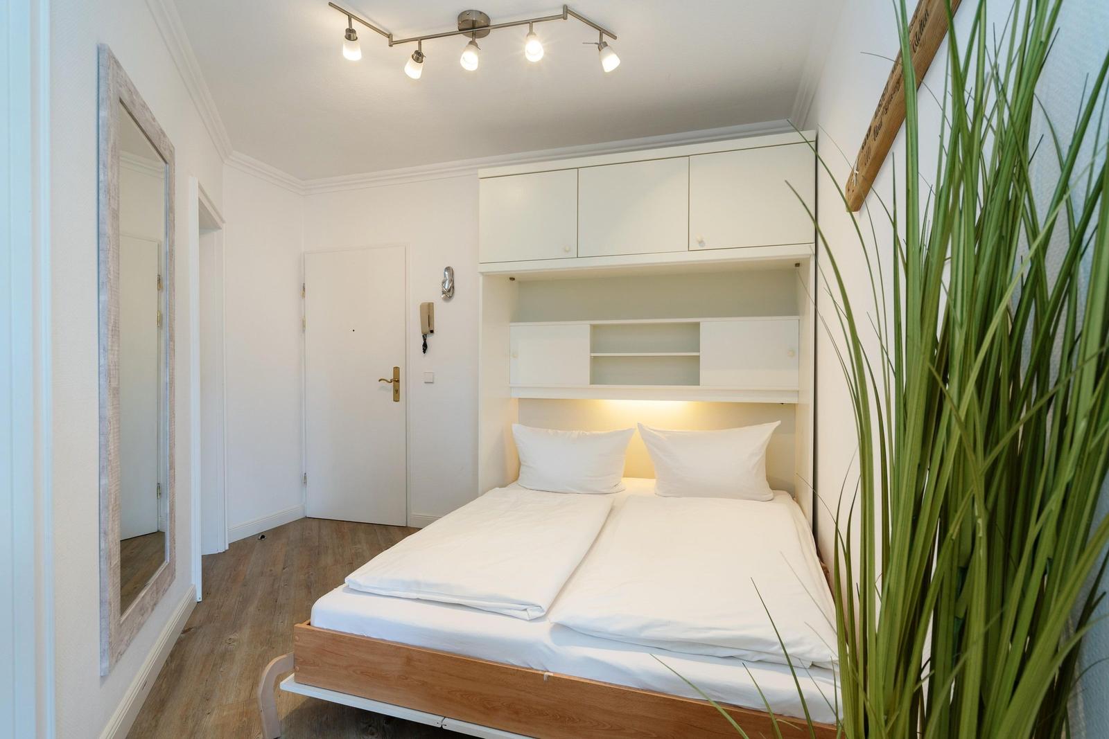 Schlafzimmer mit Doppelbett, weißen Schränken und Holzboden. Große Pflanze und Spiegel an der Wand.