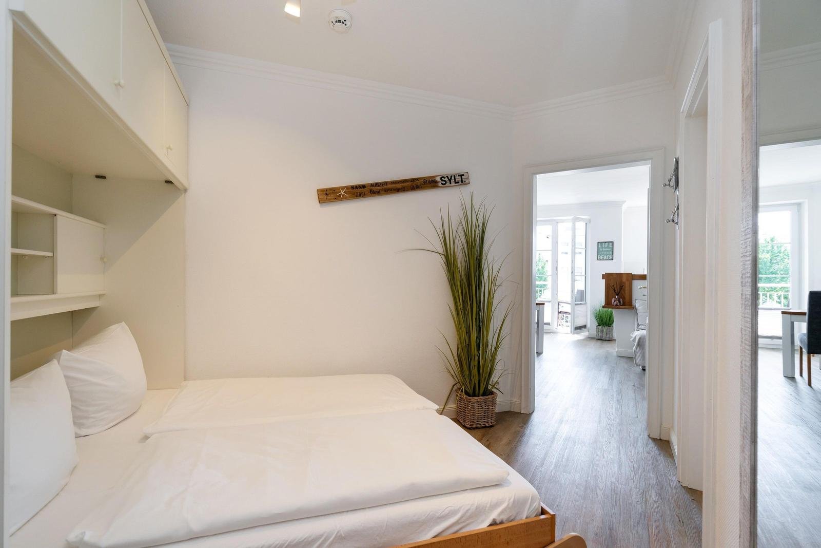 Schlafzimmer mit weißem Bett und Holzfußboden. An der Wand hängt ein hölzernes Schild mit der Aufschrift SYLT.