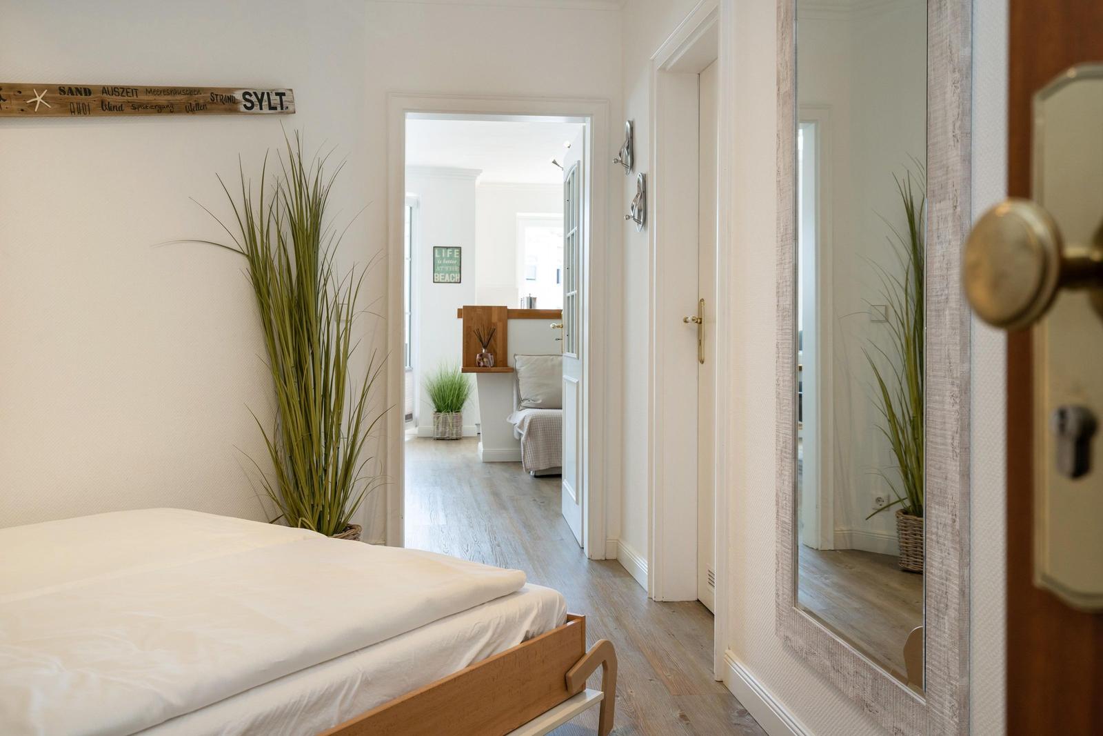 Schlafzimmer mit Bett, Pflanze und Spiegel. Durchgang zu Wohnbereich mit Sofa.