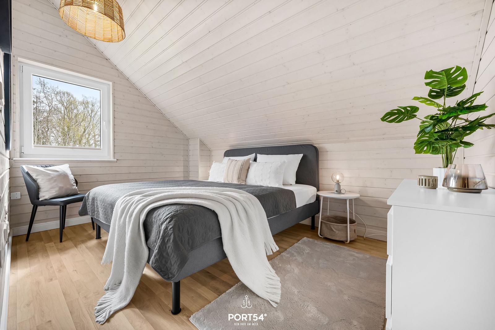 Schlafzimmer mit Bett, Nachttisch, Stuhl und Pflanze an der Wand.