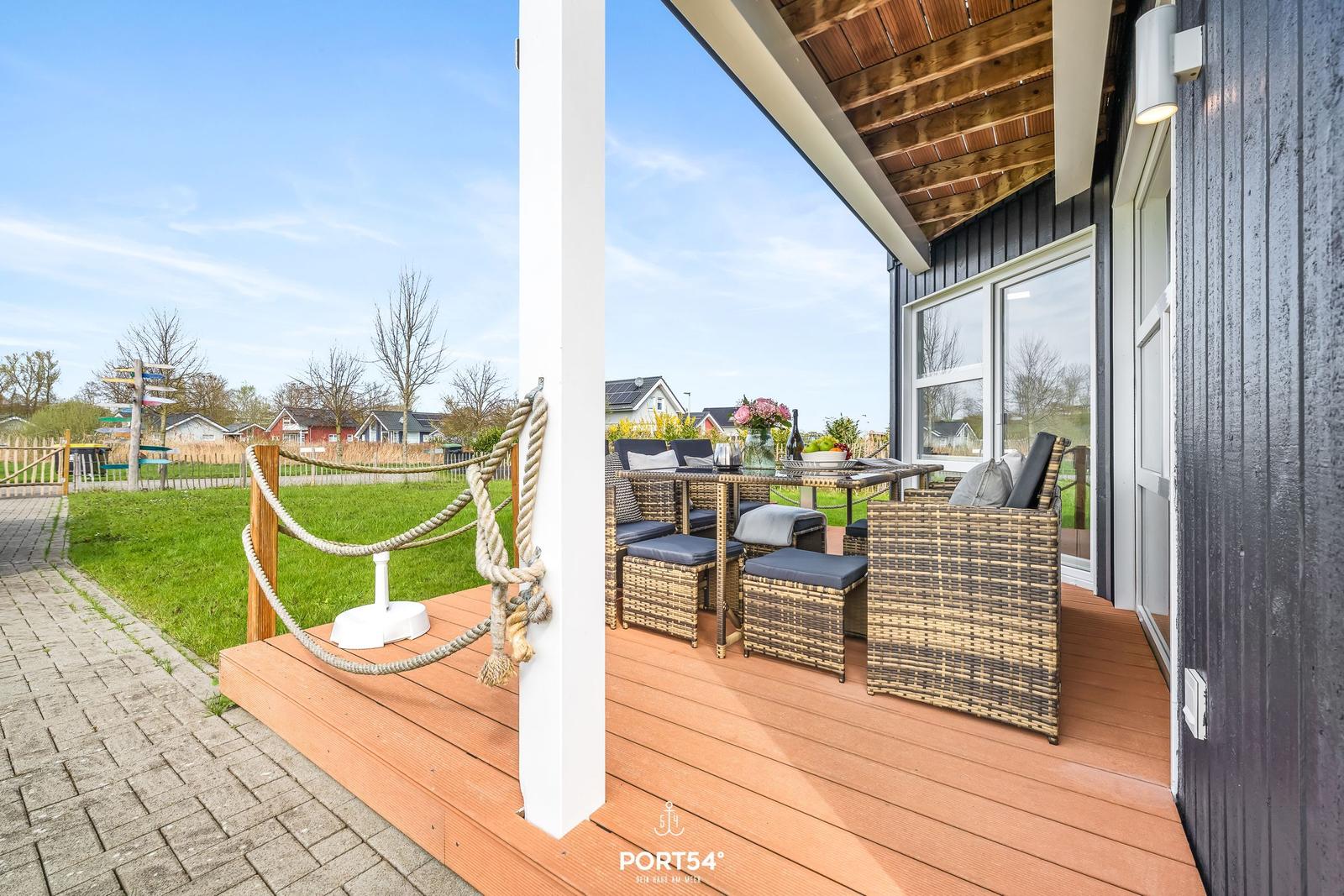 Terrasse mit Sitzgruppe und Tisch unter Holzdecke. Grünfläche und Häuser im Hintergrund.