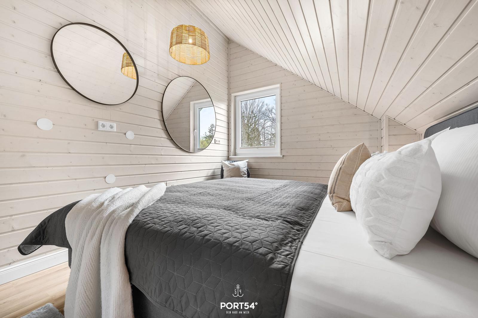 Schlafzimmer mit Bett, zwei runden Spiegeln und Fenster. Holzwand und schräges Dach.