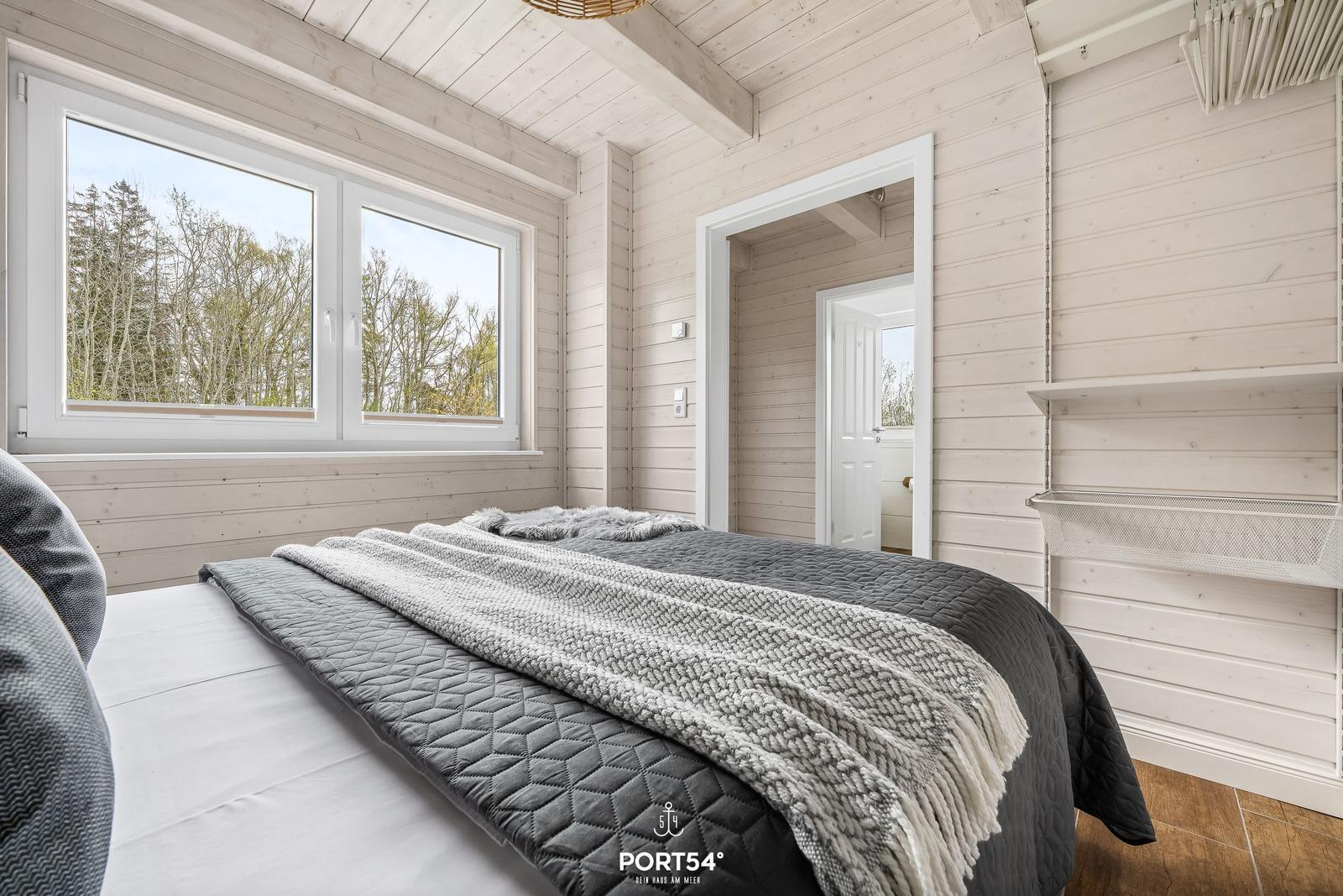Schlafzimmer mit Bett, Fenster und Holzwand. Blick auf Wald. Kleine Badezimmertür sichtbar.