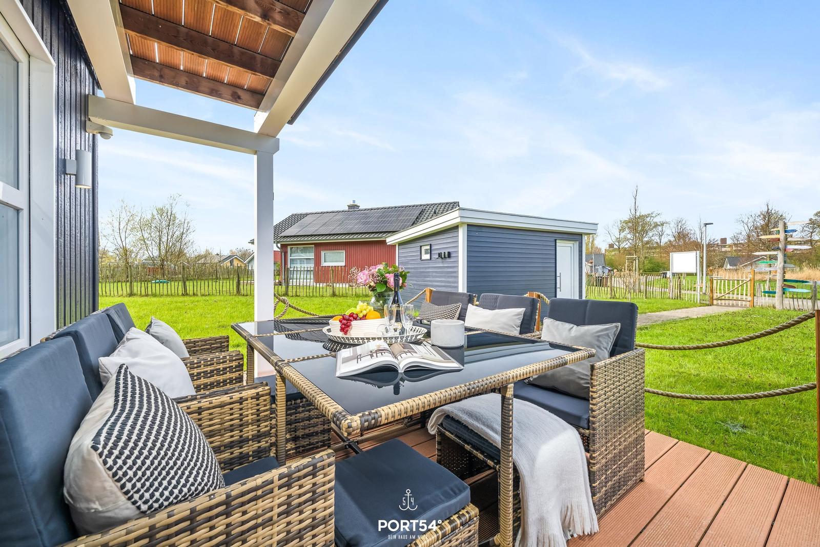Terrasse mit Sitzgruppe und Tisch unter Dach. Grünfläche und Nebengebäude im Hintergrund.