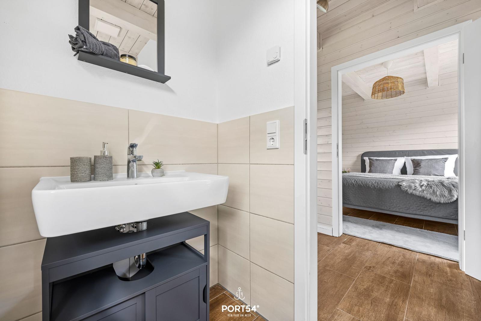 Badezimmer mit Waschbecken und Blick in das Schlafzimmer mit Bett und Decke.