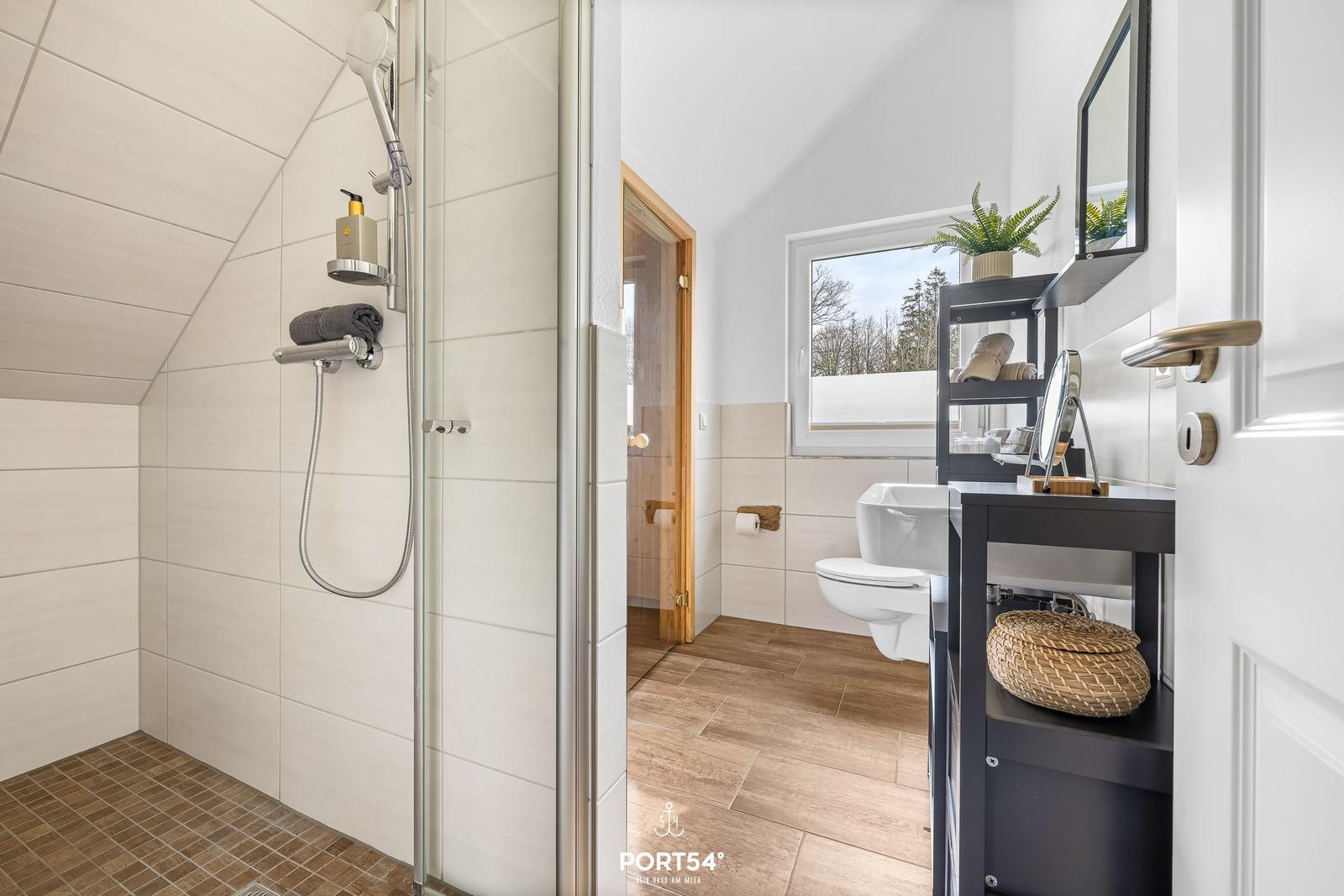 Badezimmer mit Dusche, Toilette und Waschbecken. Holzboden und Fenster mit Blick.