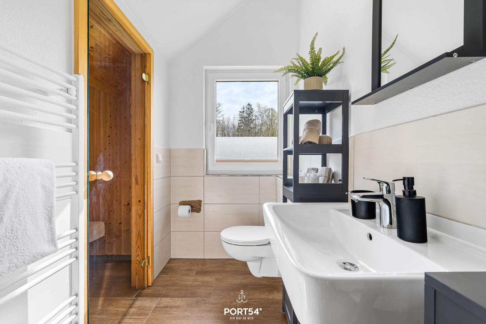 Badezimmer mit Dusche, Waschbecken und Toilette. Fenster zeigt Wald. Schwarze Regale und Pflanze.