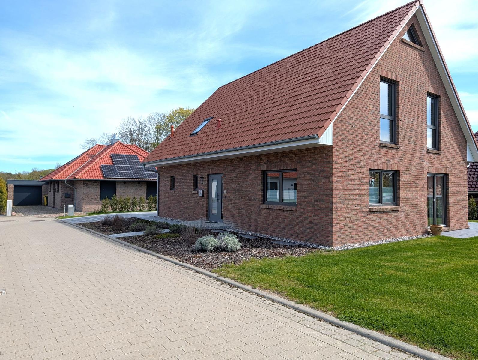 Neubauhaus mit rotem Dach und Backsteinfassade, gepflasterter Weg und Rasenflächen.