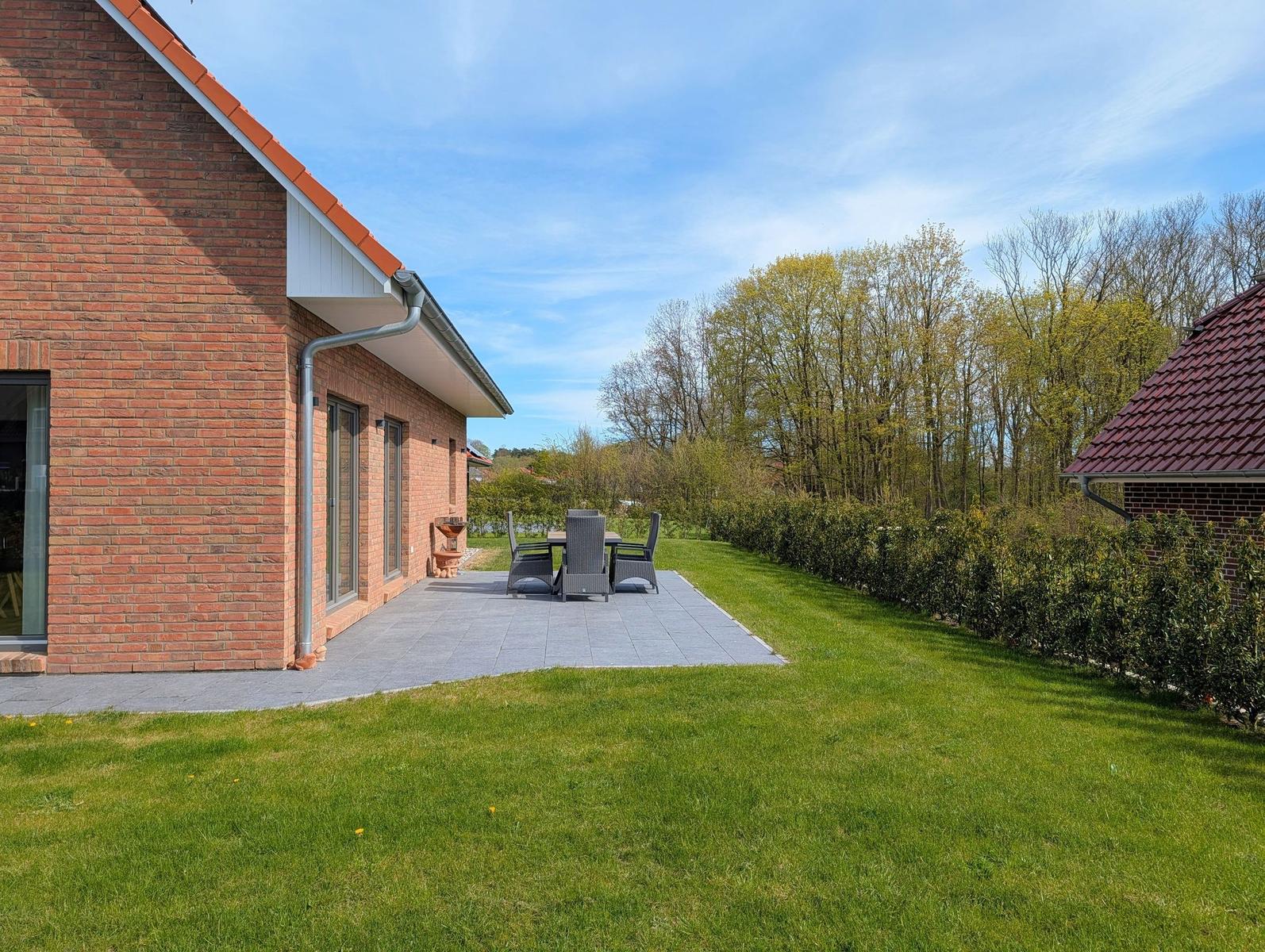 Terrasse mit Sitzgruppe neben rotem Backsteinhaus. Grasfläche und Bäume im Hintergrund.