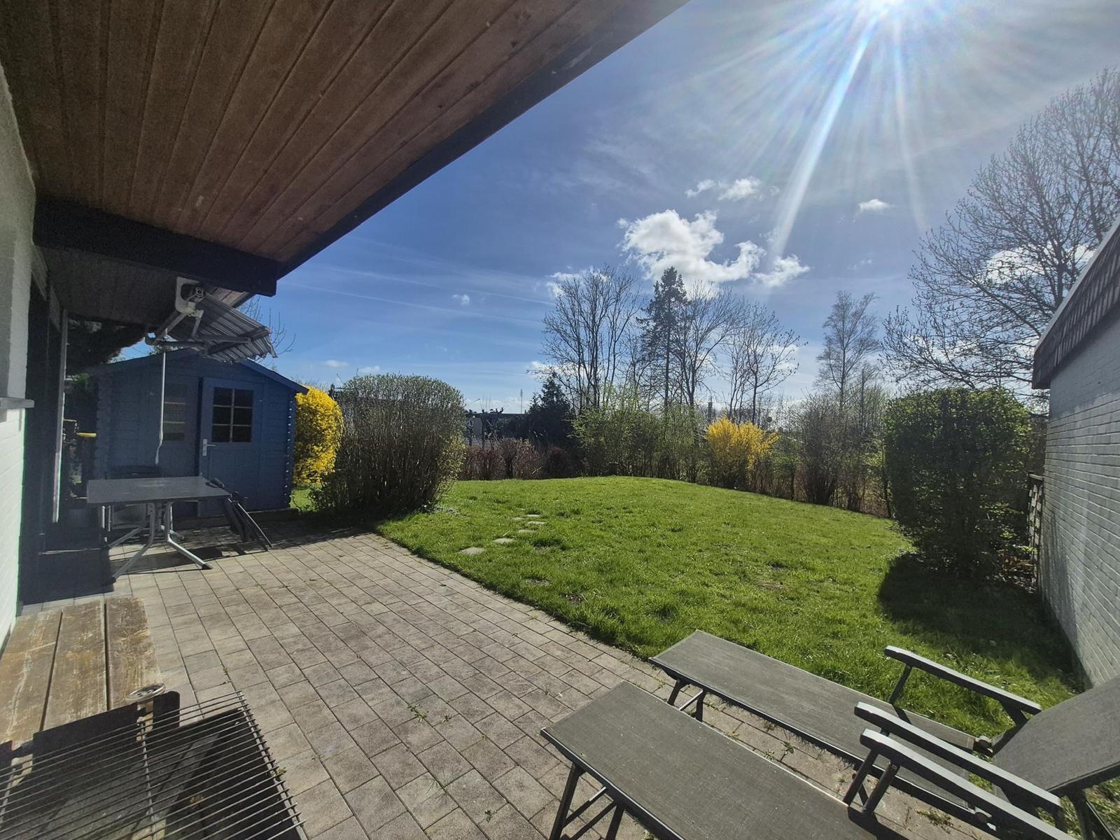 Terrace with grill, table and chairs, view of green lawn and trees under blue sky.