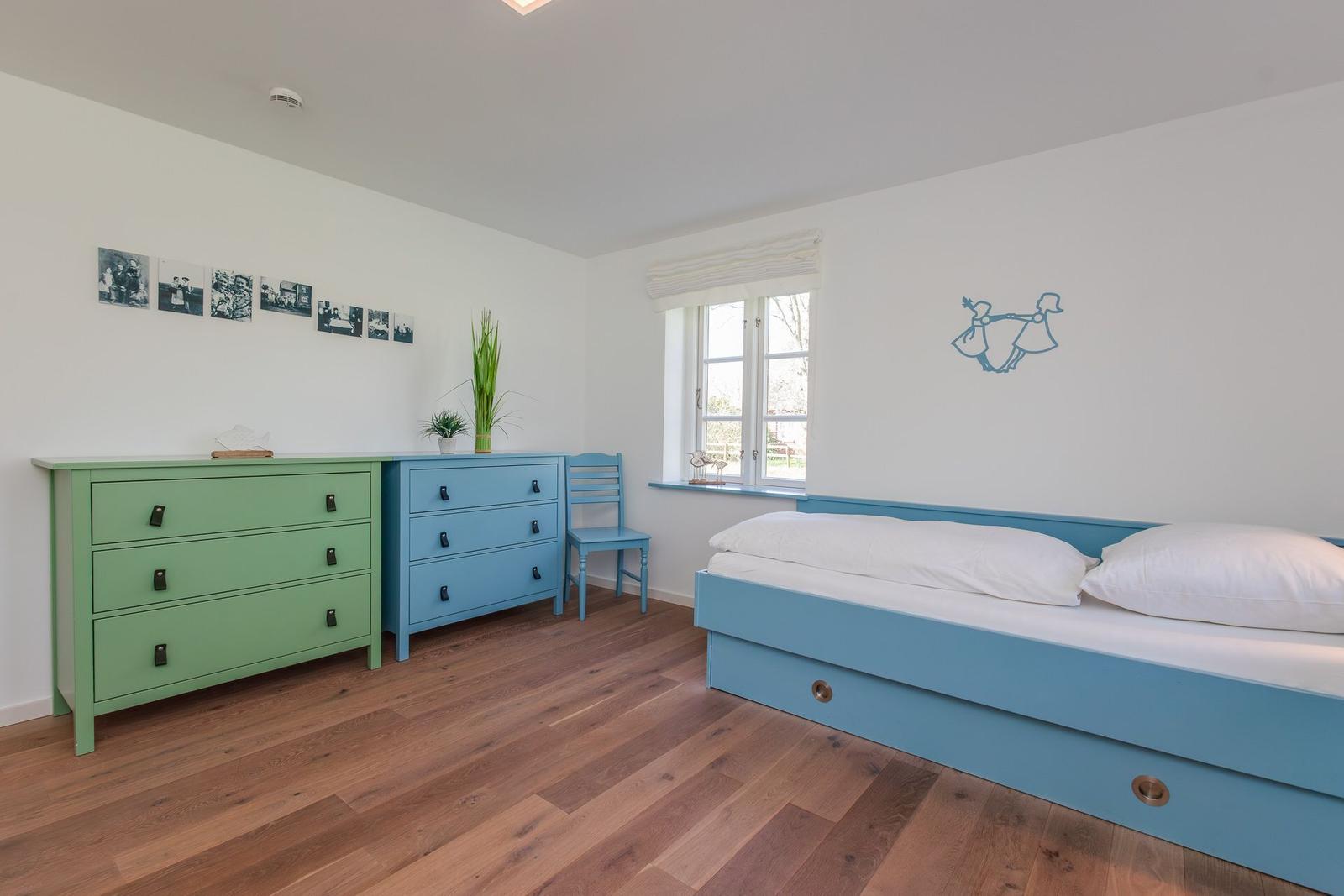 Schlafzimmer mit blauem Bett, grünem und blauem Schrank, Holzboden und Fenster mit Blick nach draußen.