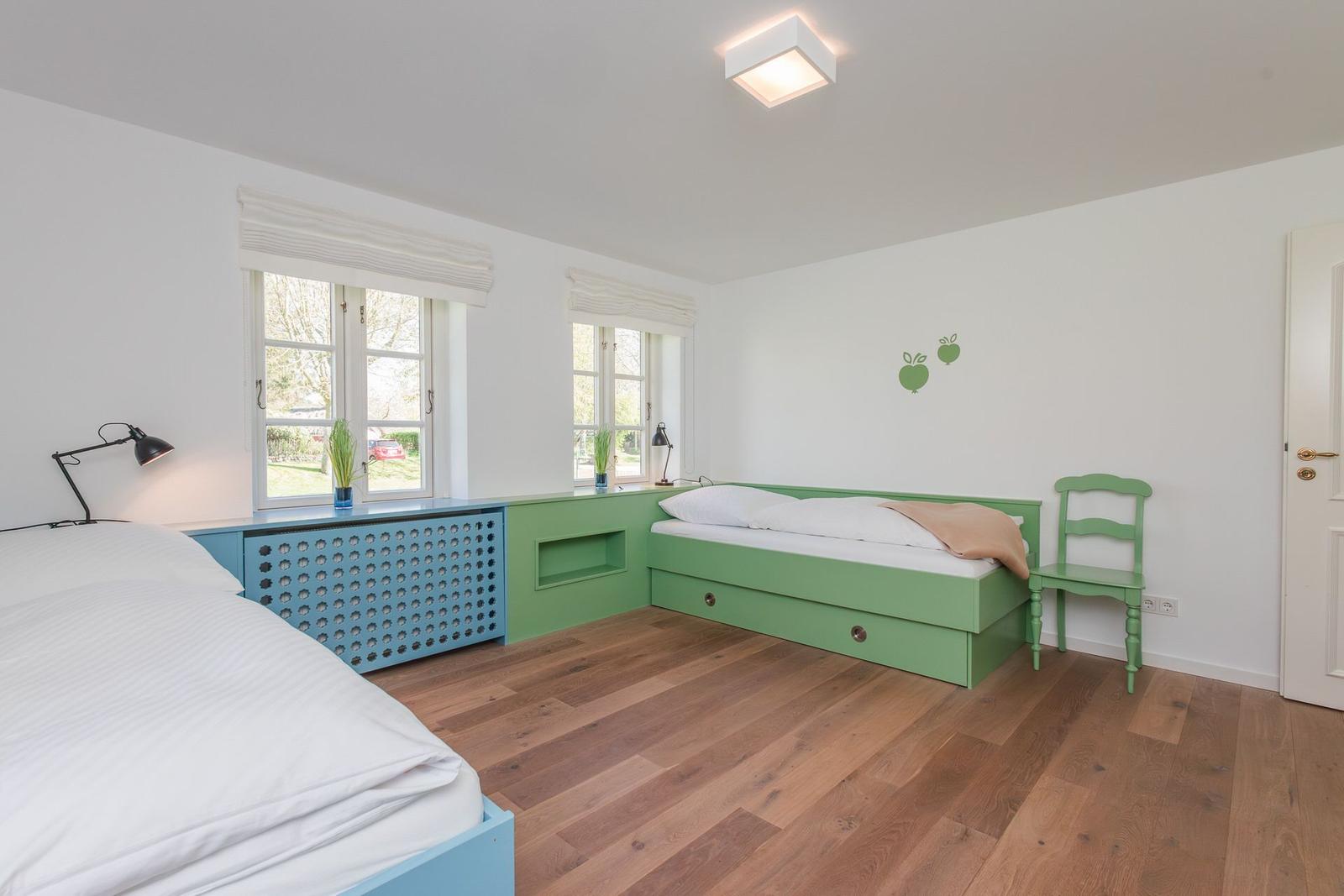 Schlafzimmer mit zwei Betten, grünen und blauen Möbeln, Holzboden und Fenstern mit Blick nach draußen.