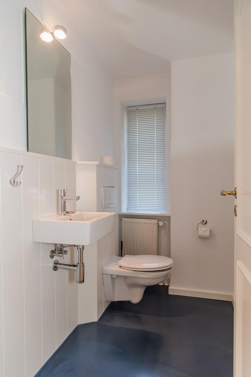 Badezimmer mit Waschbecken, Spiegel, WC und Fenster mit Jalousie.