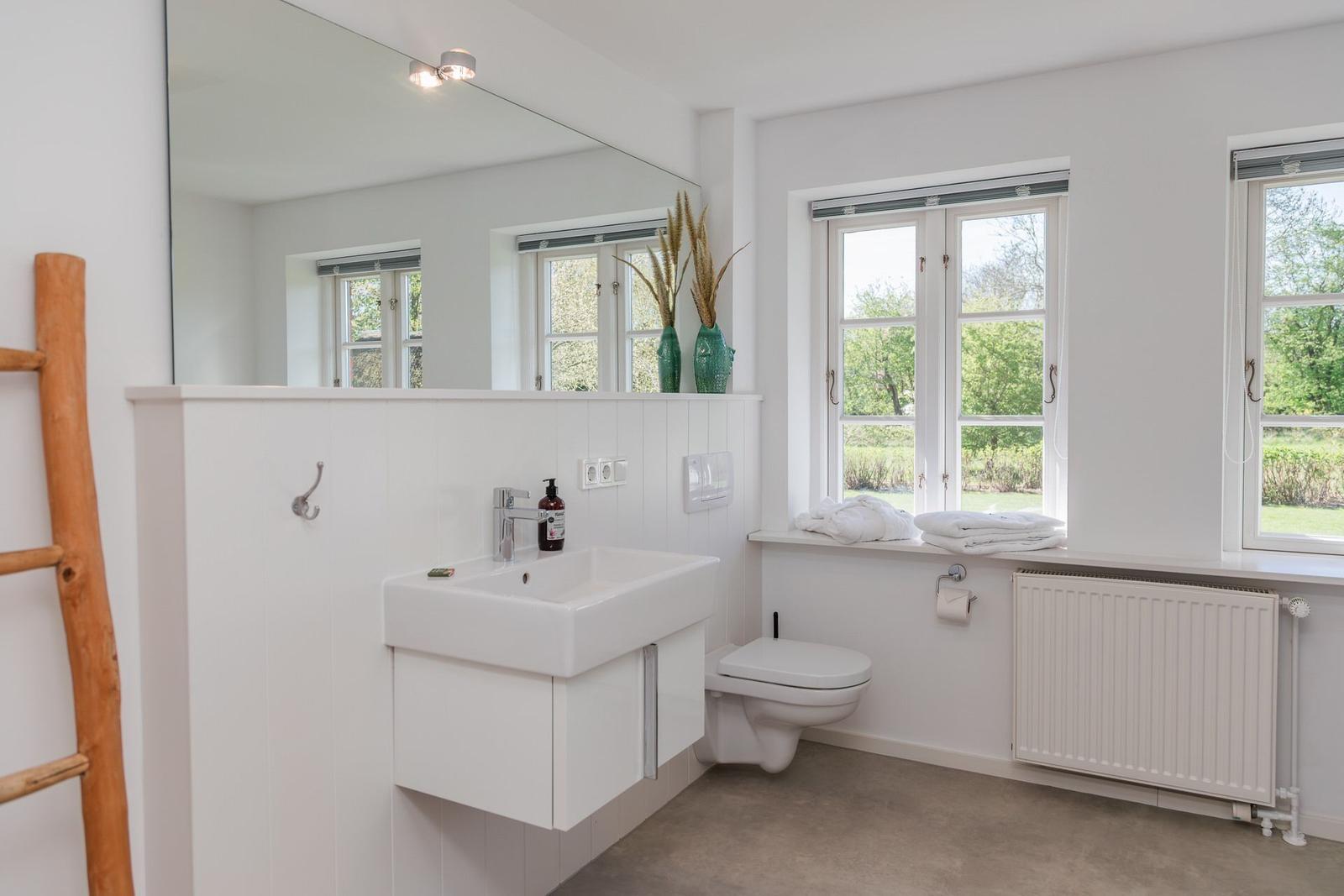 Badezimmer mit Waschbecken, Spiegel, Toilette und Fenstern mit Blick auf Grün.