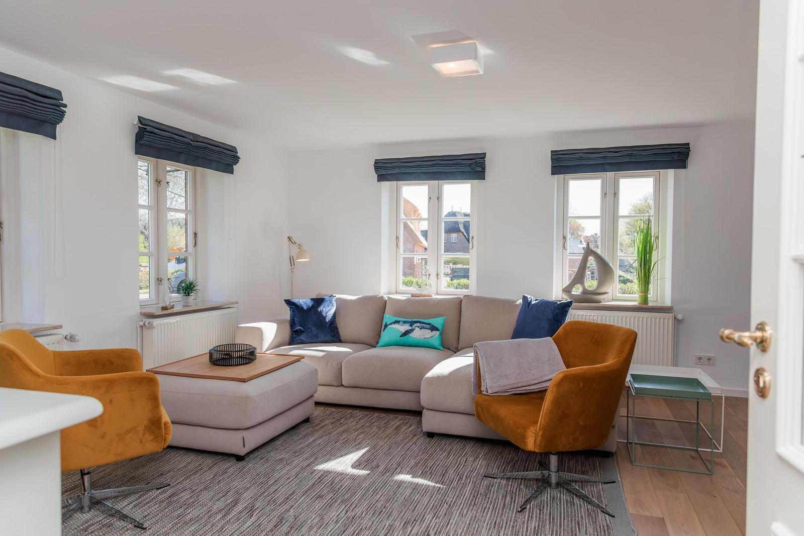 Wohnzimmer mit Sofa, zwei orangefarbenen Sesseln und Fenstern mit dunklen Vorhängen.