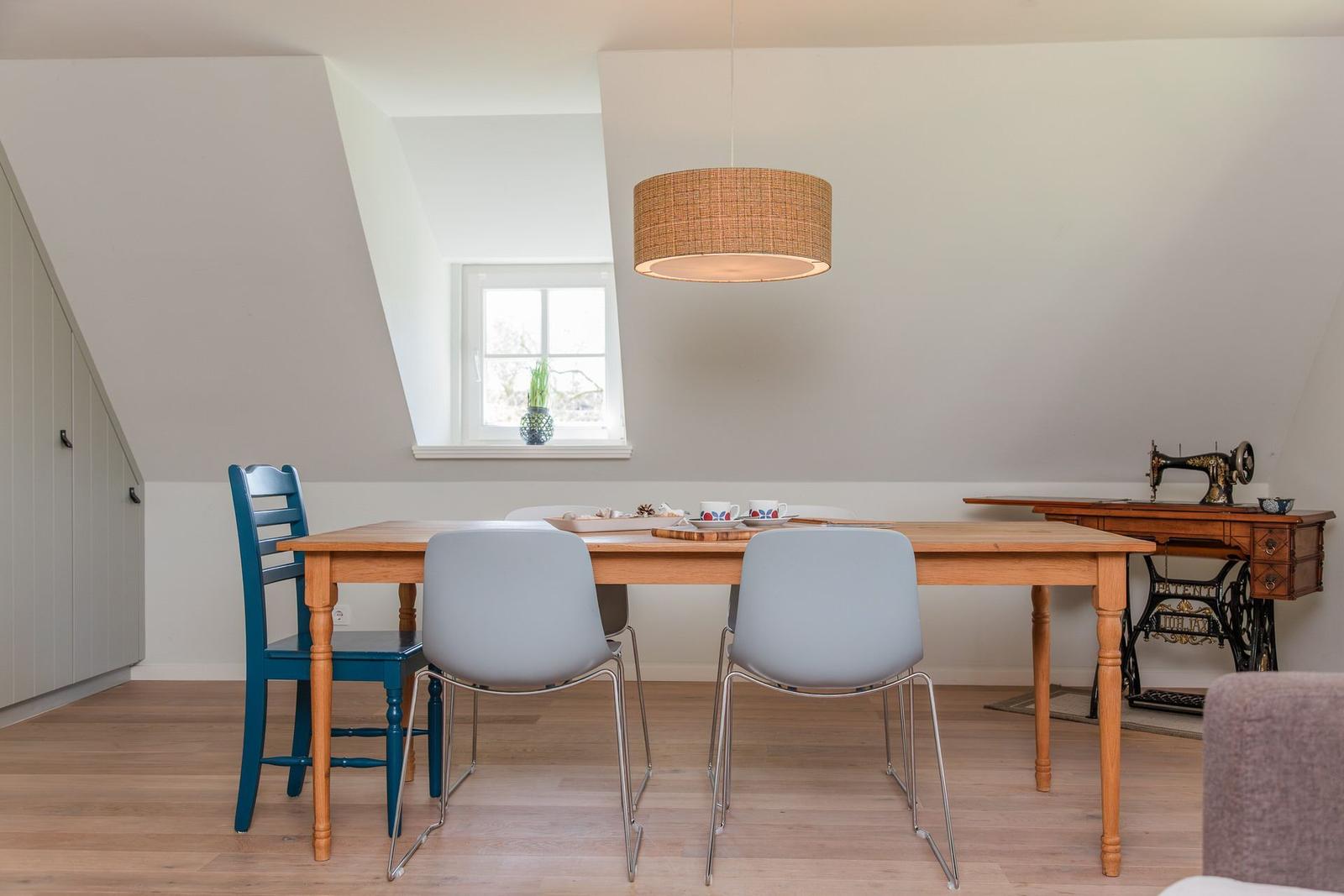 Esszimmer mit Holztisch, Stühlen und Fenster. An der Wand steht eine Nähmaschine.