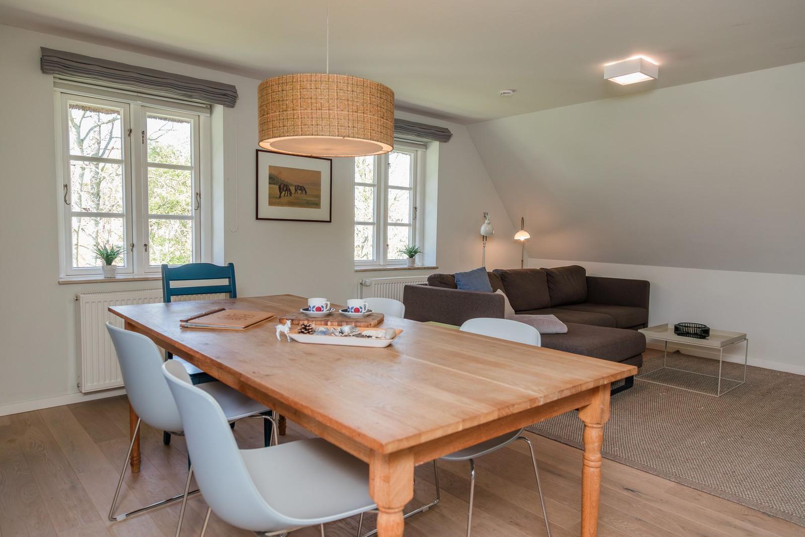 Ein Essbereich mit Holztisch und Stühlen, daneben ein Wohnbereich mit Sofa und Fenstern.