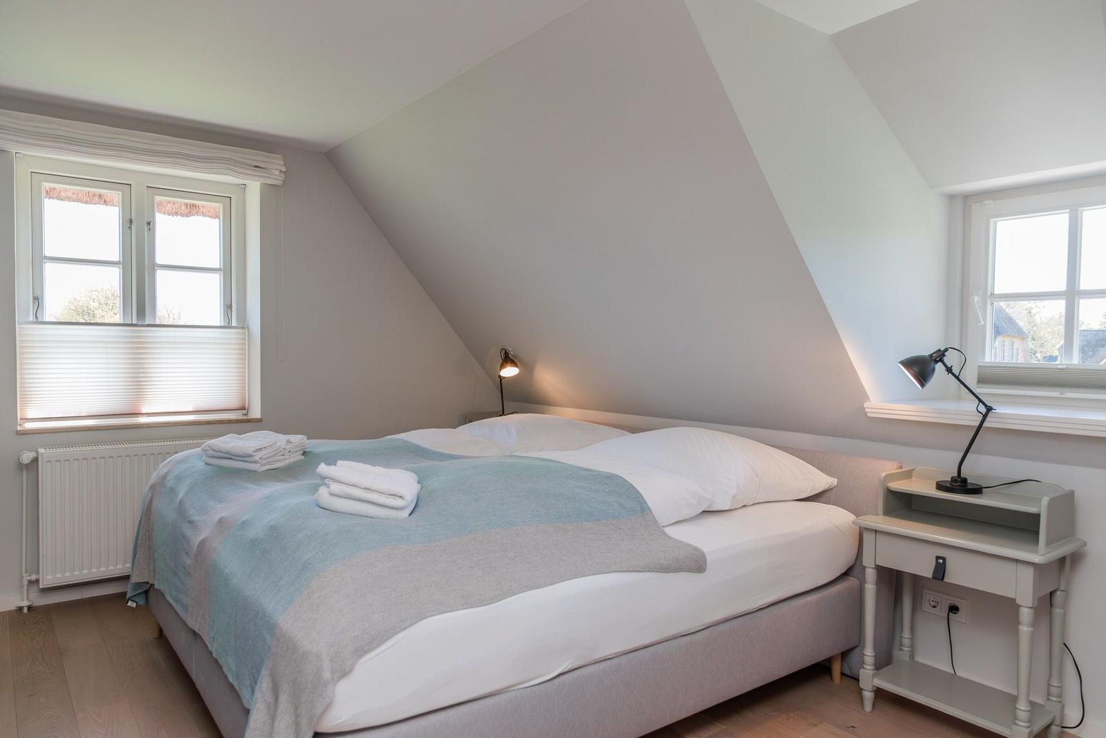 Dachgeschosszimmer mit Bett, zwei Fenstern und Nachttisch mit Lampe.