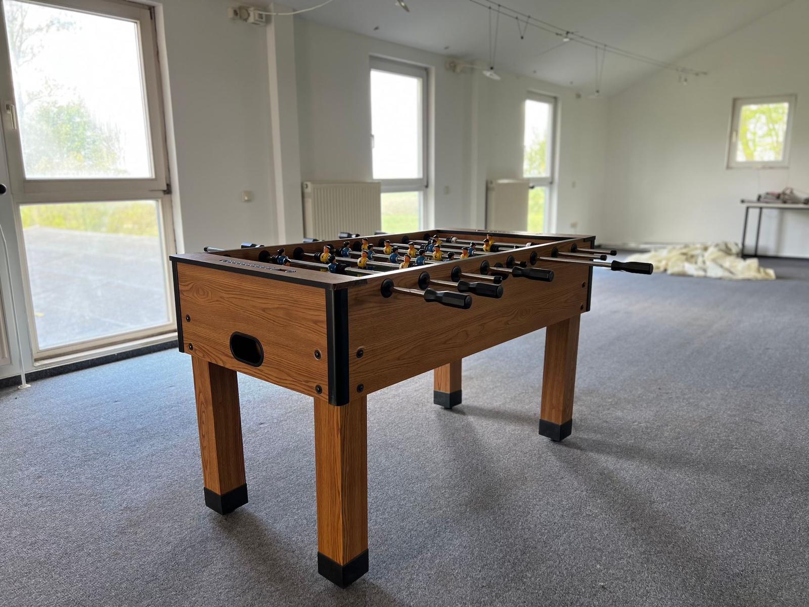 Fußballtisch aus Holz in einem hellen Raum mit Fenstern und Teppichboden.