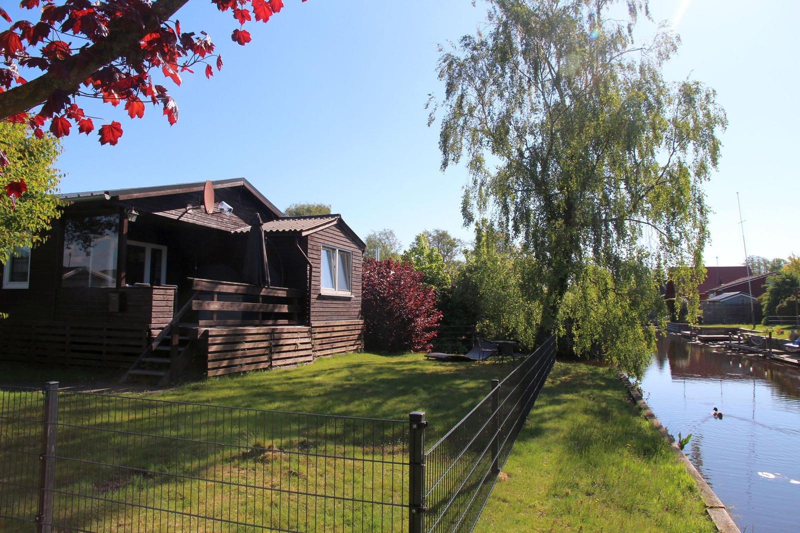 Wooden house with terrace, garden, and canal. Trees and red leaves in the foreground.