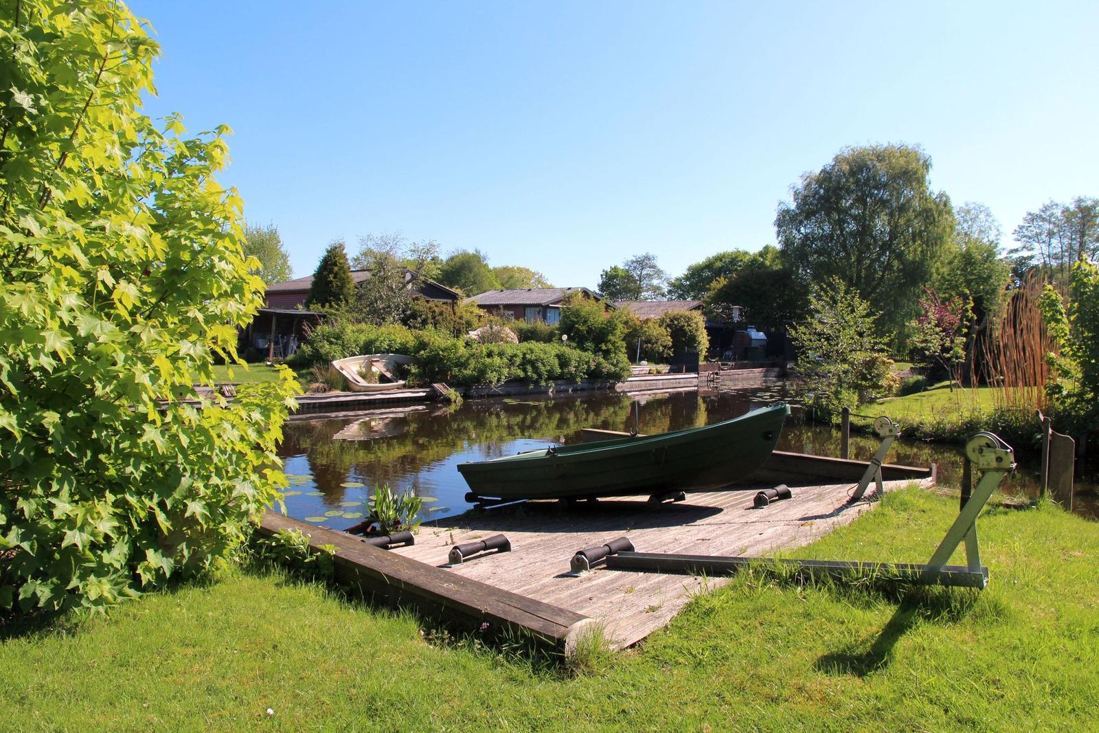 A green boat rests on a wooden platform by a lake. Houses and trees are visible in the background.
