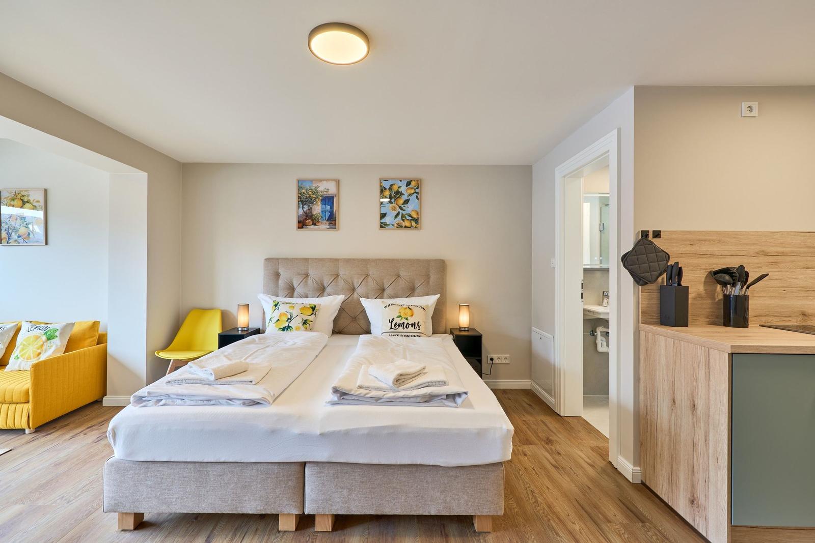 Schlafzimmer mit Doppelbett, gelber Sitzgelegenheit und Küchenbereich.
