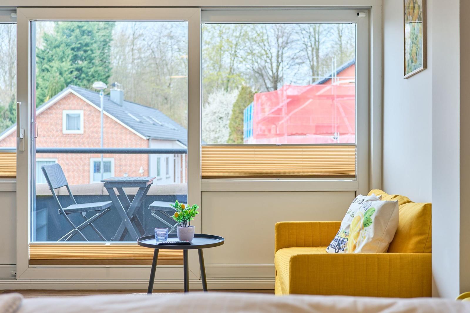 Ein gelbes Sofa mit Kissen steht vor einem Fenster mit Balkon. Draußen ist ein Tisch und Stühle zu sehen.