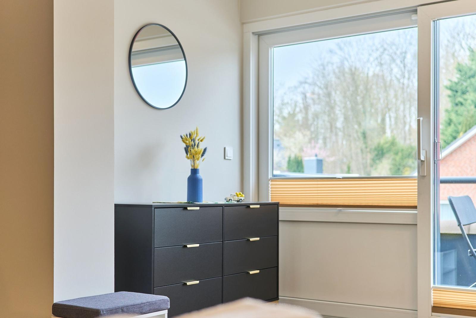 Schwarzer Schrank mit blauem Vasen und runden Spiegel an der Wand. Fenster mit Blick auf Balkon.