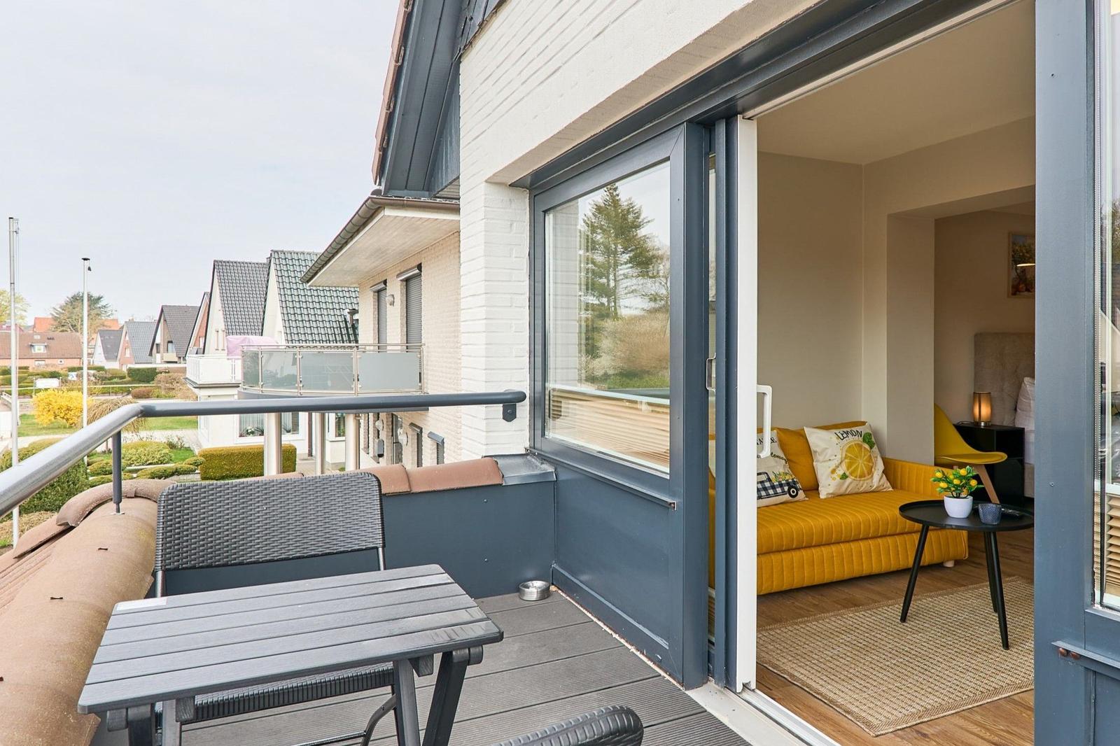 Terrasse mit Tisch und Stuhl. Durch Glastür ist Wohnzimmer mit gelbem Sofa sichtbar.