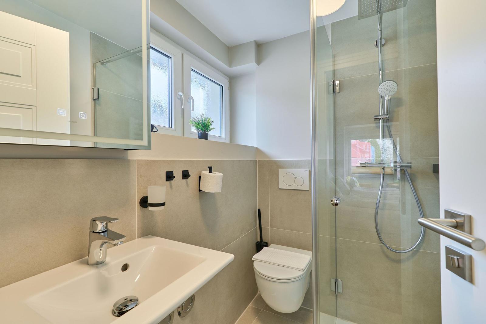 Badezimmer mit Dusche, Toilette und Waschbecken unter Fenster.