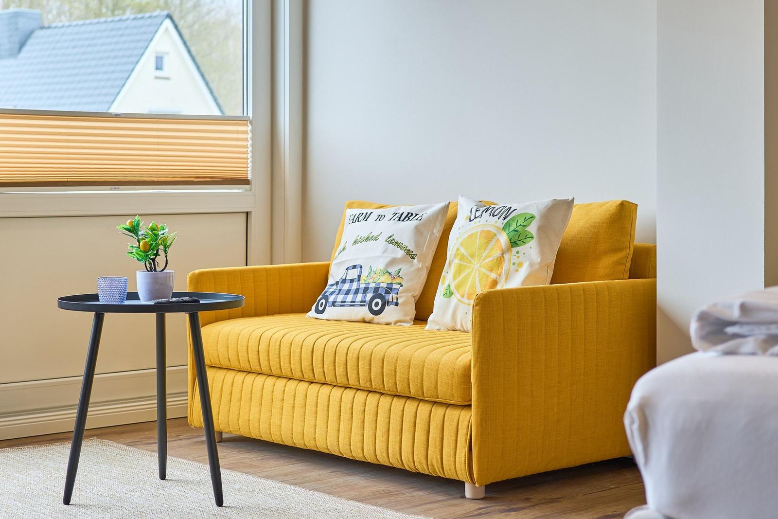 Gelber Sessel mit Kissen, kleiner Tisch und Pflanze vor Fenster.