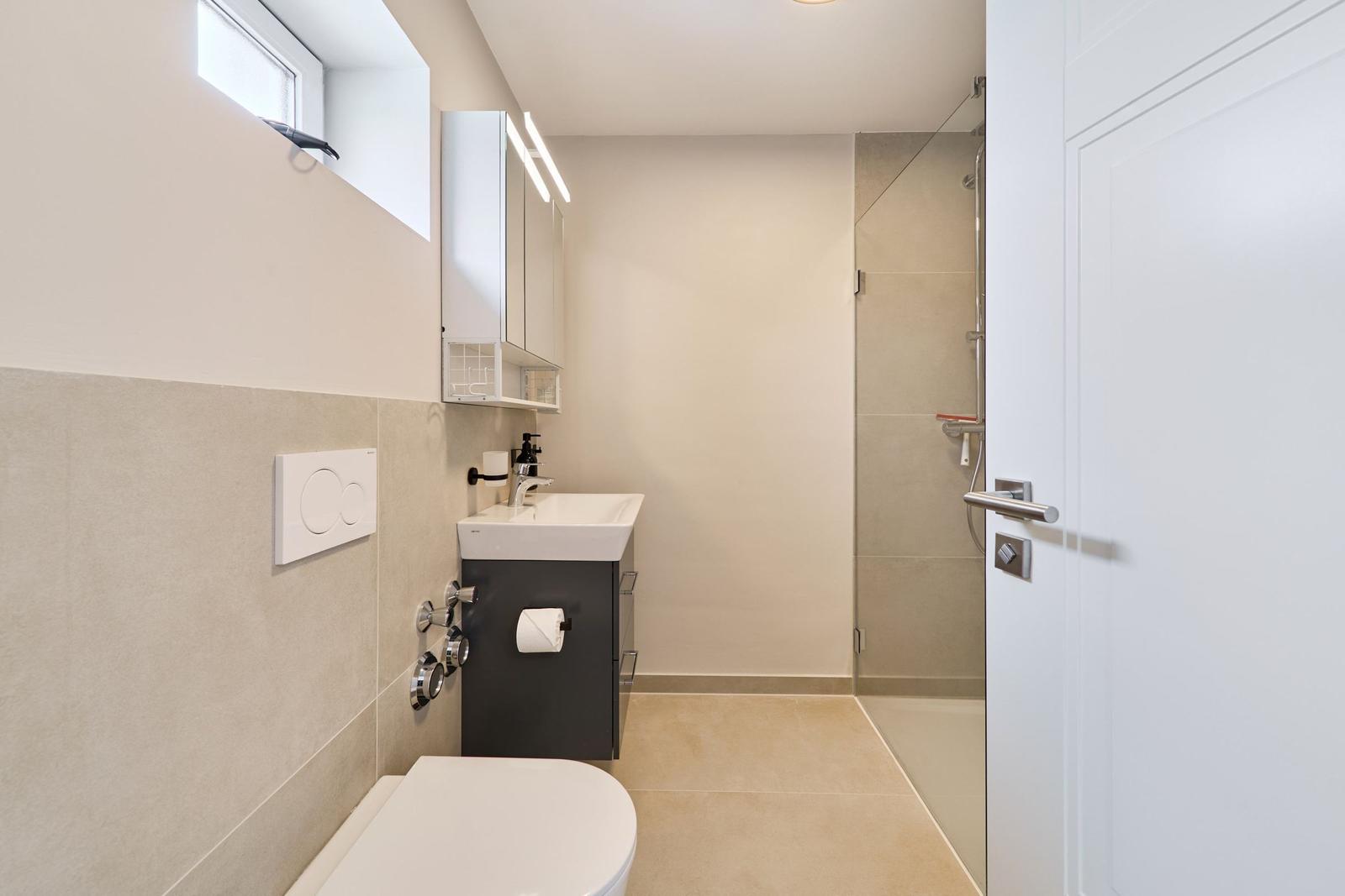 Badezimmer mit Dusche, Waschbecken und Toilette. Helle Wandfarbe und beigefarbene Fliesen.