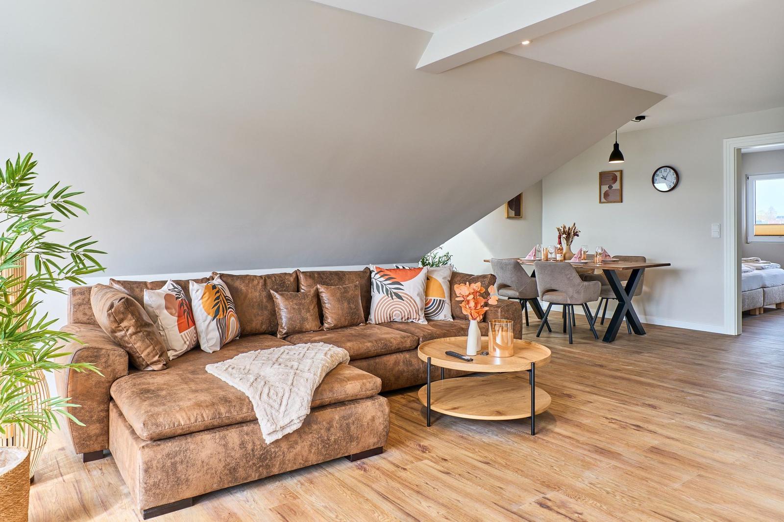 Wohnbereich mit braunem Sofa, Essbereich und Holzboden. Dachschrägen und Pflanzen sichtbar.