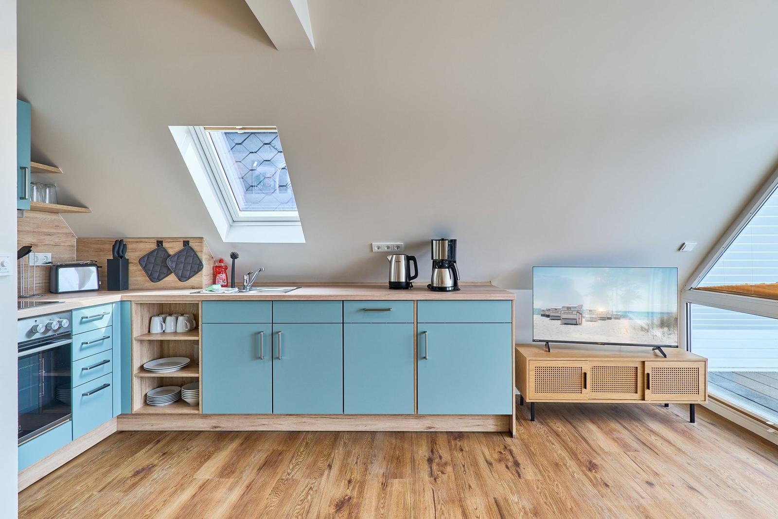 Küche mit blauen Schränken und Holzoberfläche. Fernseher auf Holzregal. Dachfenster und großes Fenster.