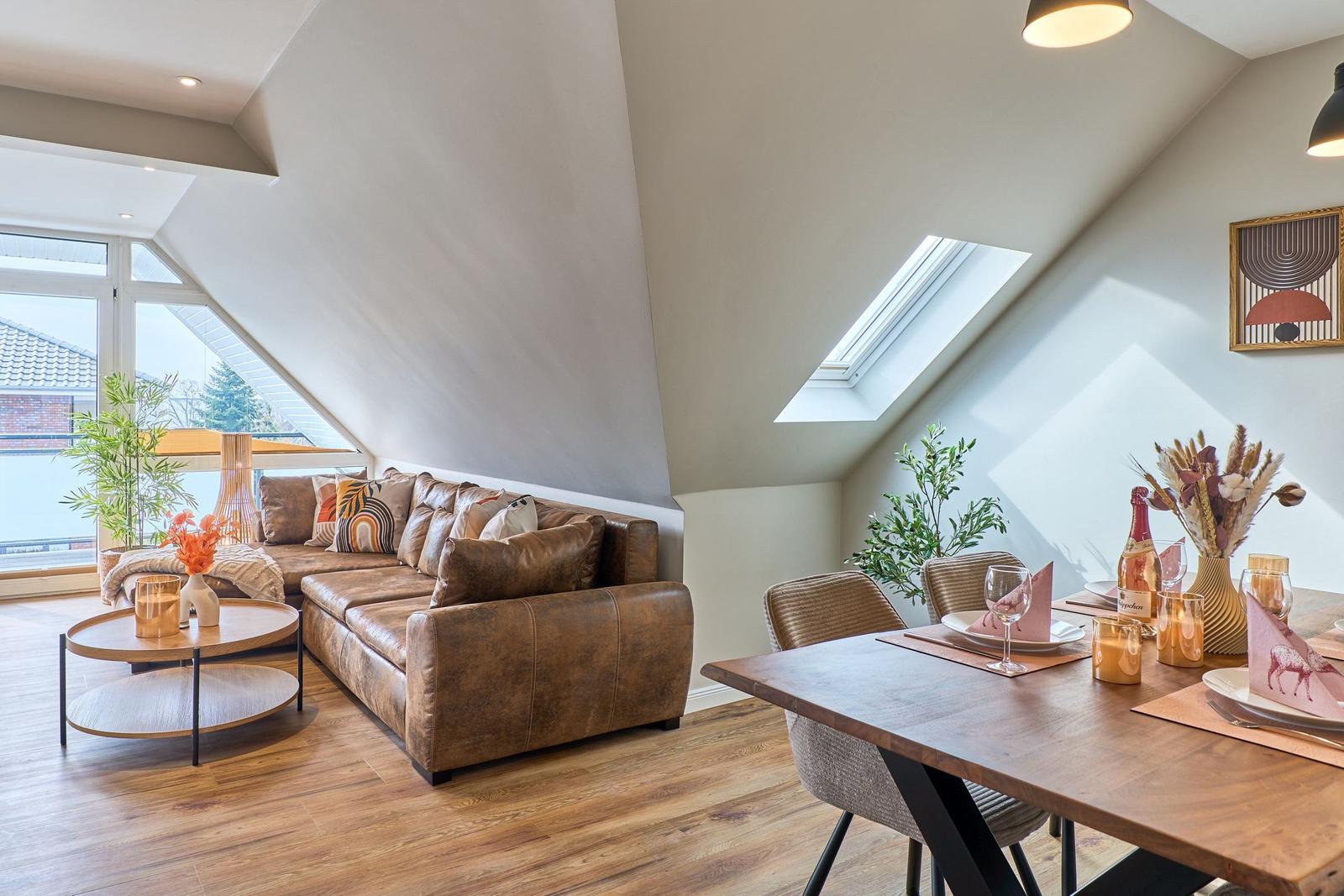 Wohn- und Essbereich mit Sofa, Tisch und großen Fenstern. Dachfenster und Holzboden.