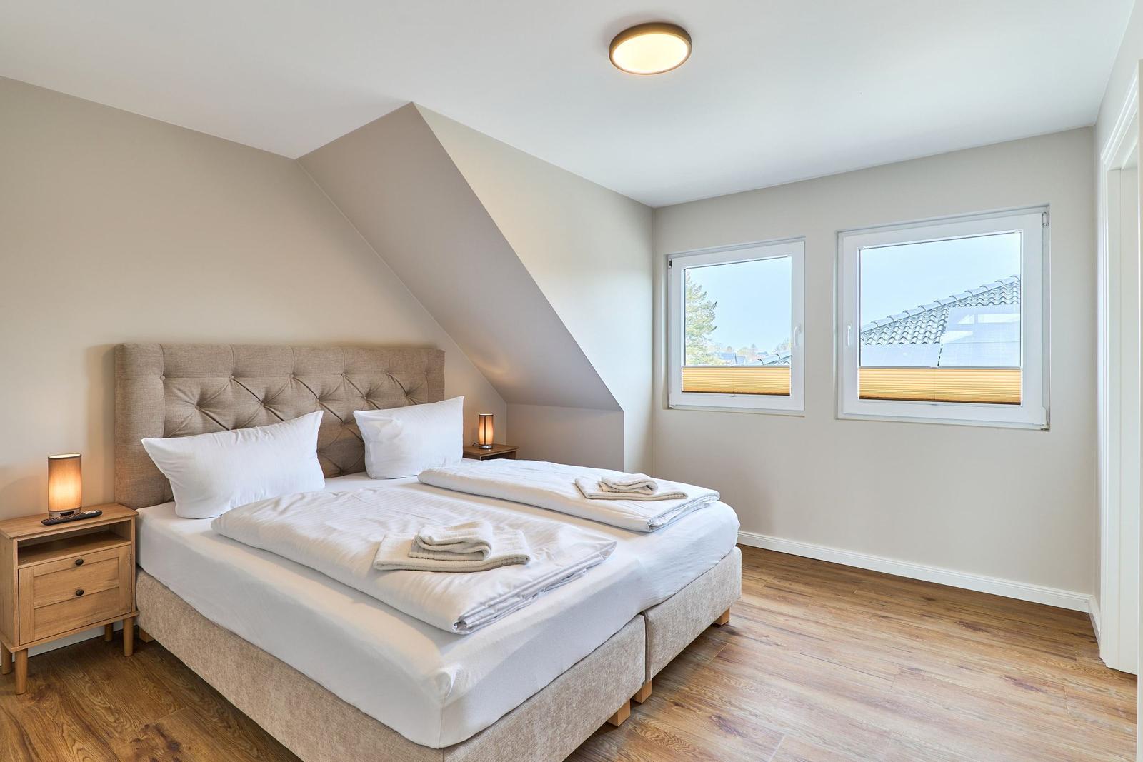 Schlafzimmer mit Doppelbett, zwei Fenstern und Holzboden.