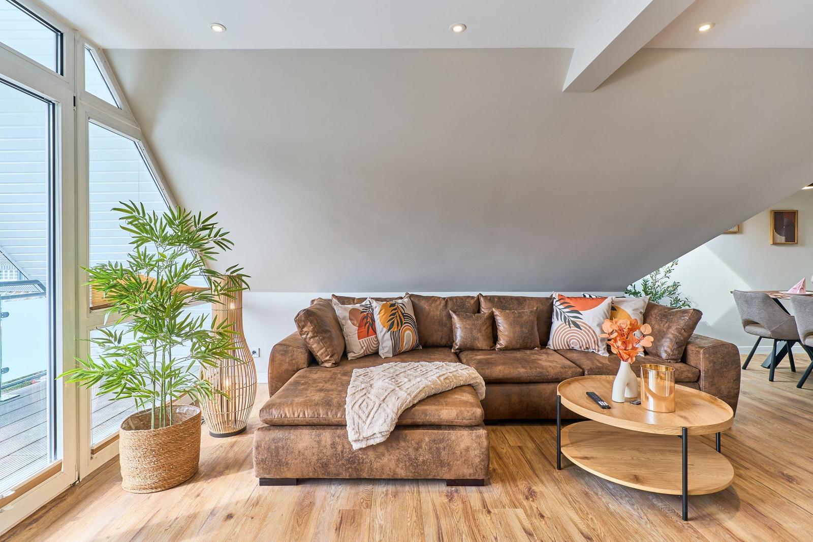 Ein Wohnzimmer mit braunem Sofa, Holzboden und großen Fenstern.
