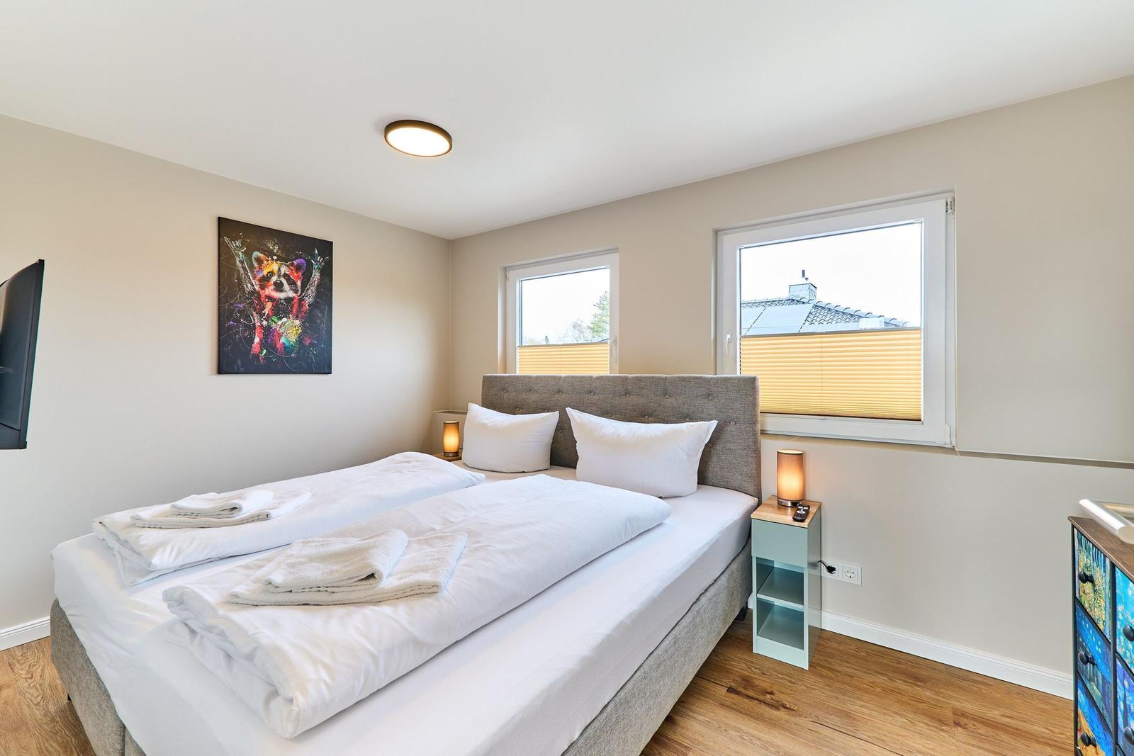 Schlafzimmer mit Doppelbett, Fenster und Wandbild. Holzboden und Nachttisch mit Lampe.