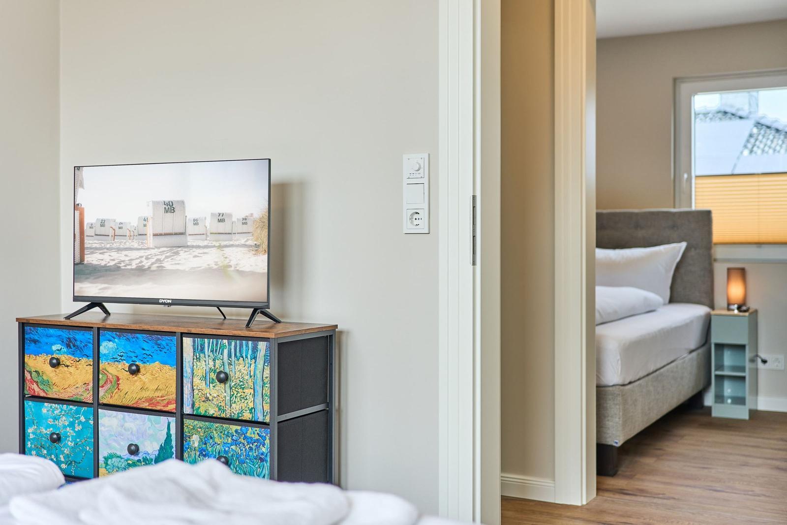 TV auf Schrank mit Van Goghs Motiven. Blick in Schlafzimmer mit Bett und Nachttisch.