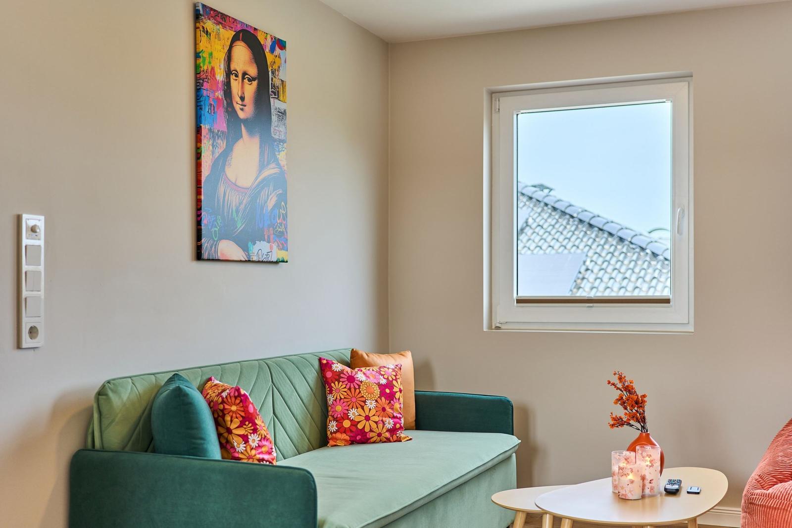 Grünes Sofa mit Kissen, Wandbild und Fenster mit Blick auf Dach.