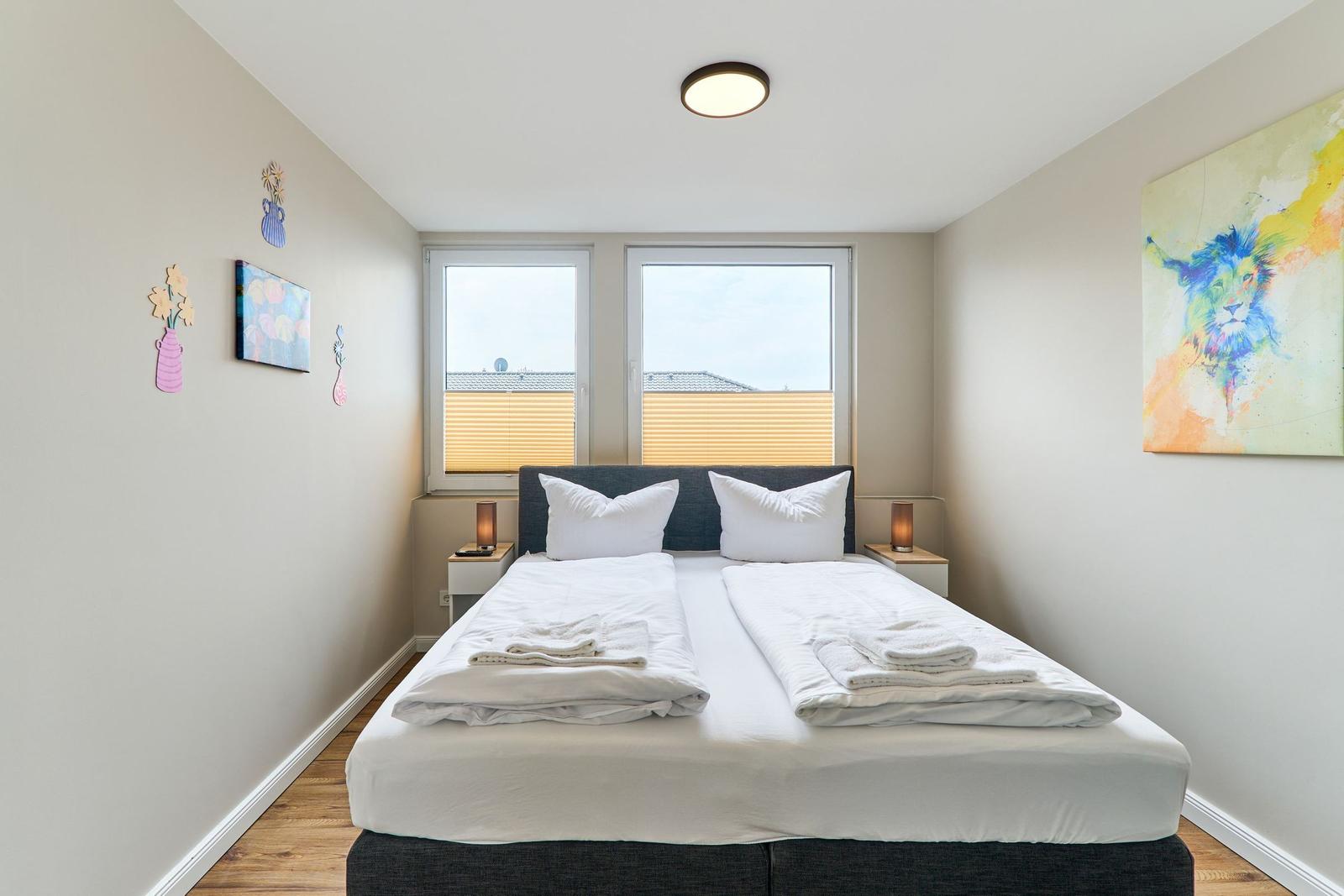 Schlafzimmer mit Doppelbett, Fenstern und Wandbildern.