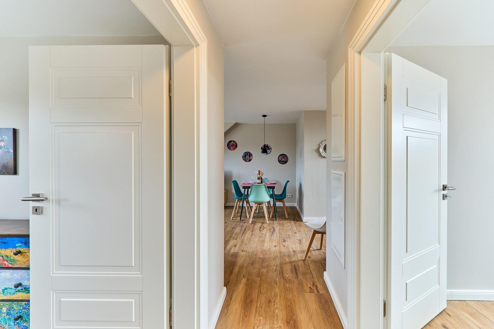 Durchgang mit weißen Türen führt in ein Esszimmer mit Holzboden und bunten Stühlen.